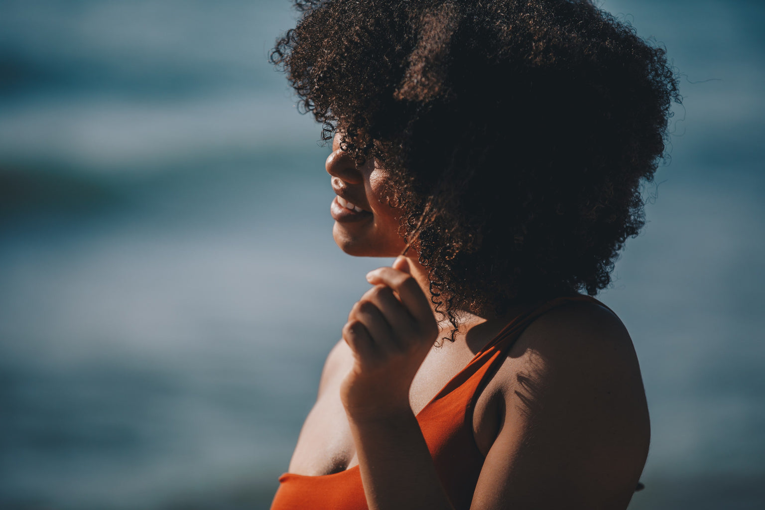 Woman with an afro looking out at water, wearing an orange top.